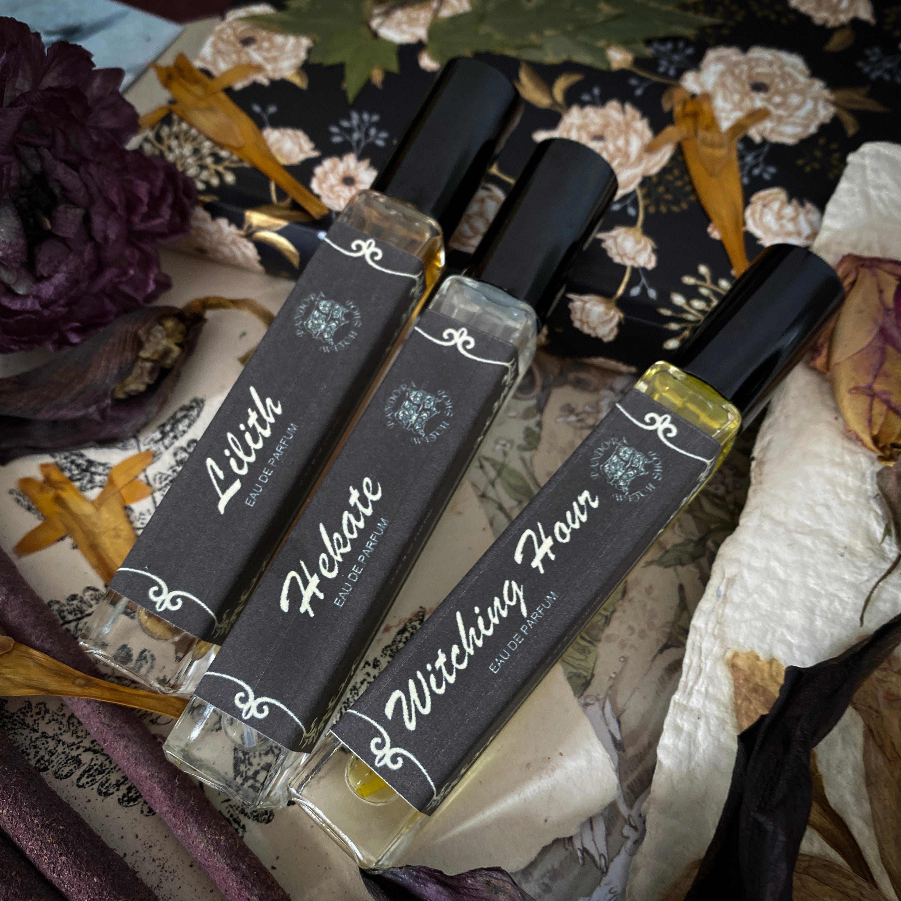 Three black glass bottles of ritual perfumes labeled Lilith, Witching Hour, and Hekate. Styled with dried flowers, candles, and witchcraft accessories on a dark mystical background. Product photo for a witch shop with international shipping