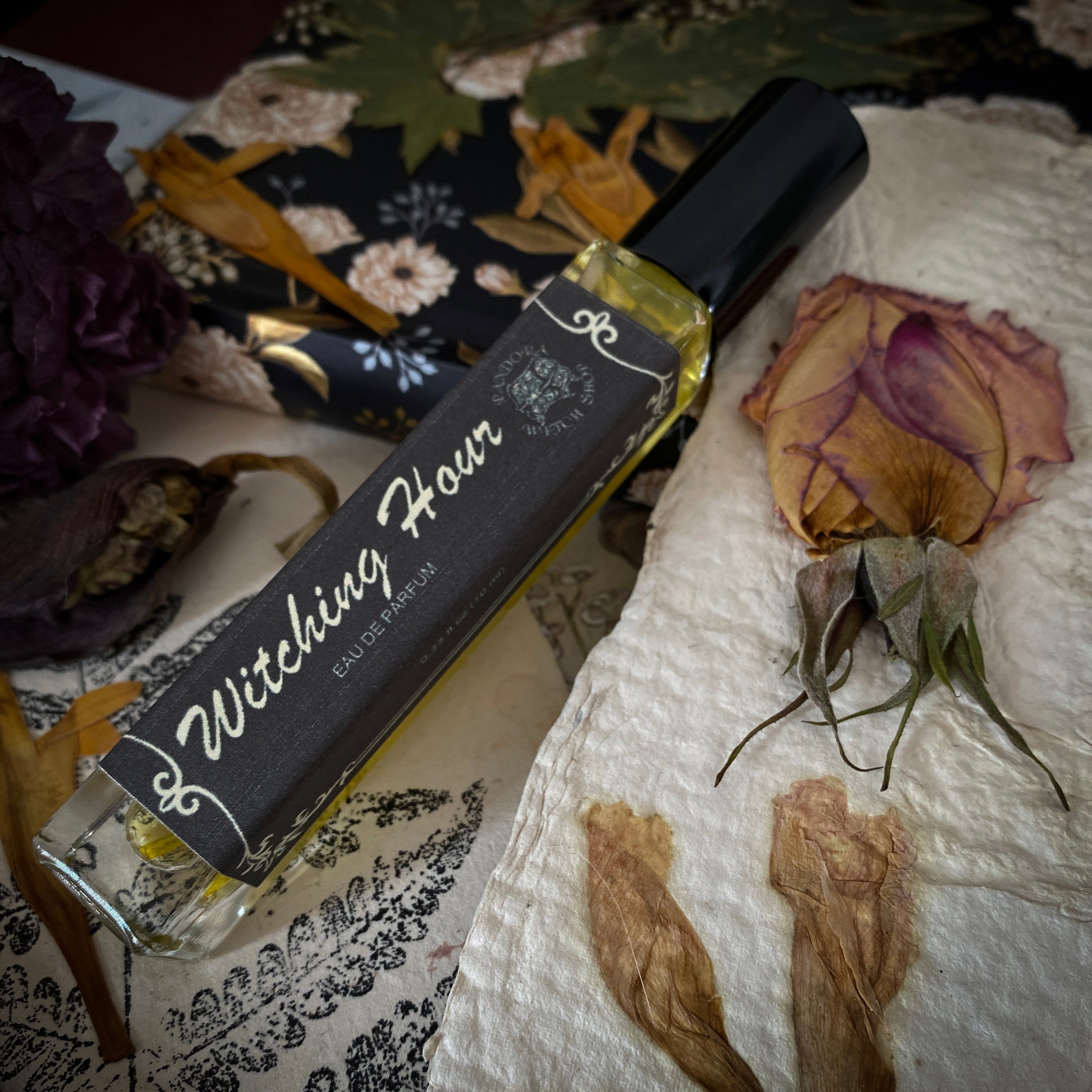 Perfume bottle labeled 'Witching Hour' on a textured surface with dried flowers.