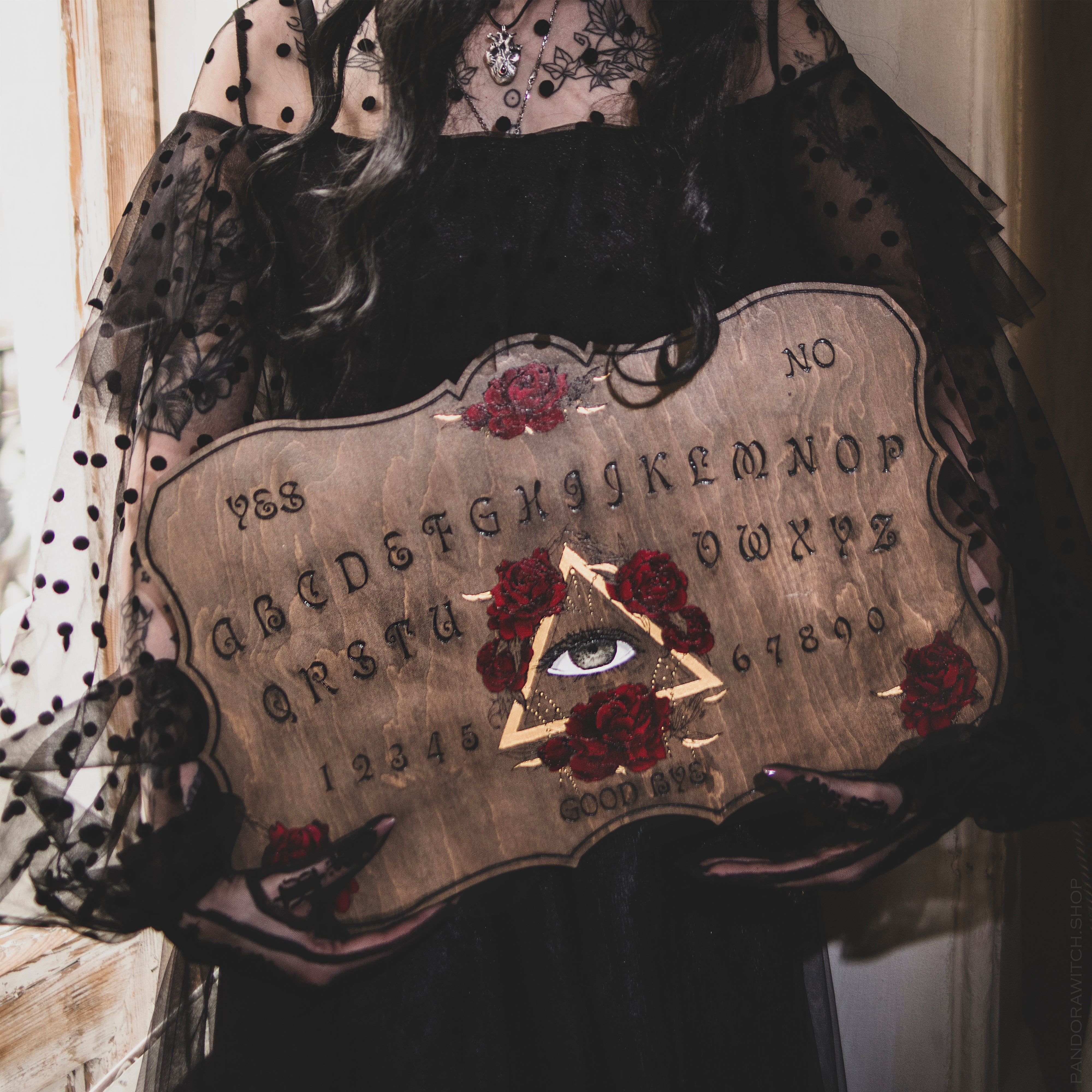A person holding a wooden Ouija board with the All Seeing Eye and blooming roses engraved on it, dressed in a black, floral-patterned dress.