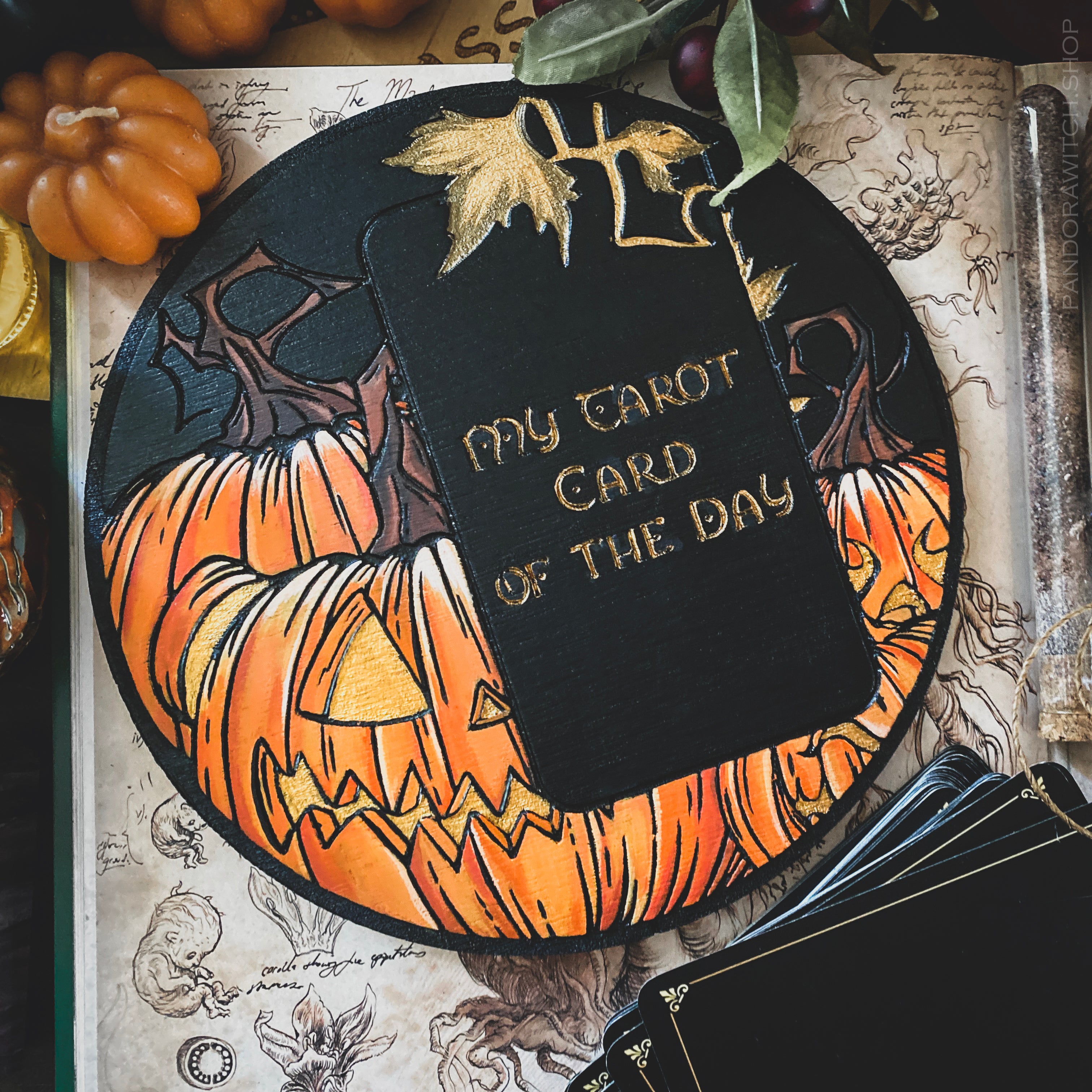 A circular wooden board with a colorful engraving of pumpkins and leaves, with a space for a tarot card, placed on a table with autumn-themed decorations.