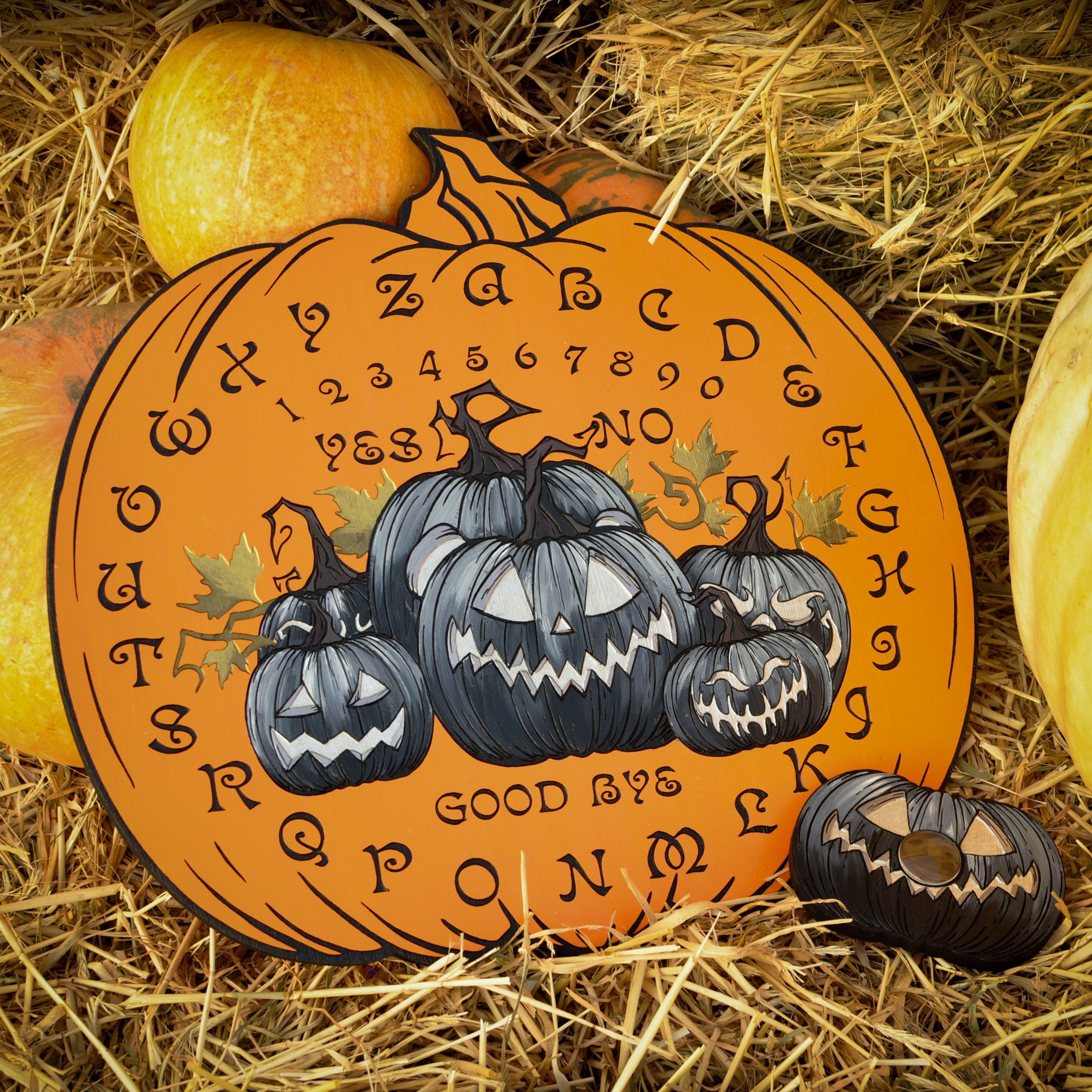 An orange Ouija board with pumpkin designs and letters, placed on a hay bale with pumpkins and autumn leaves in the background.