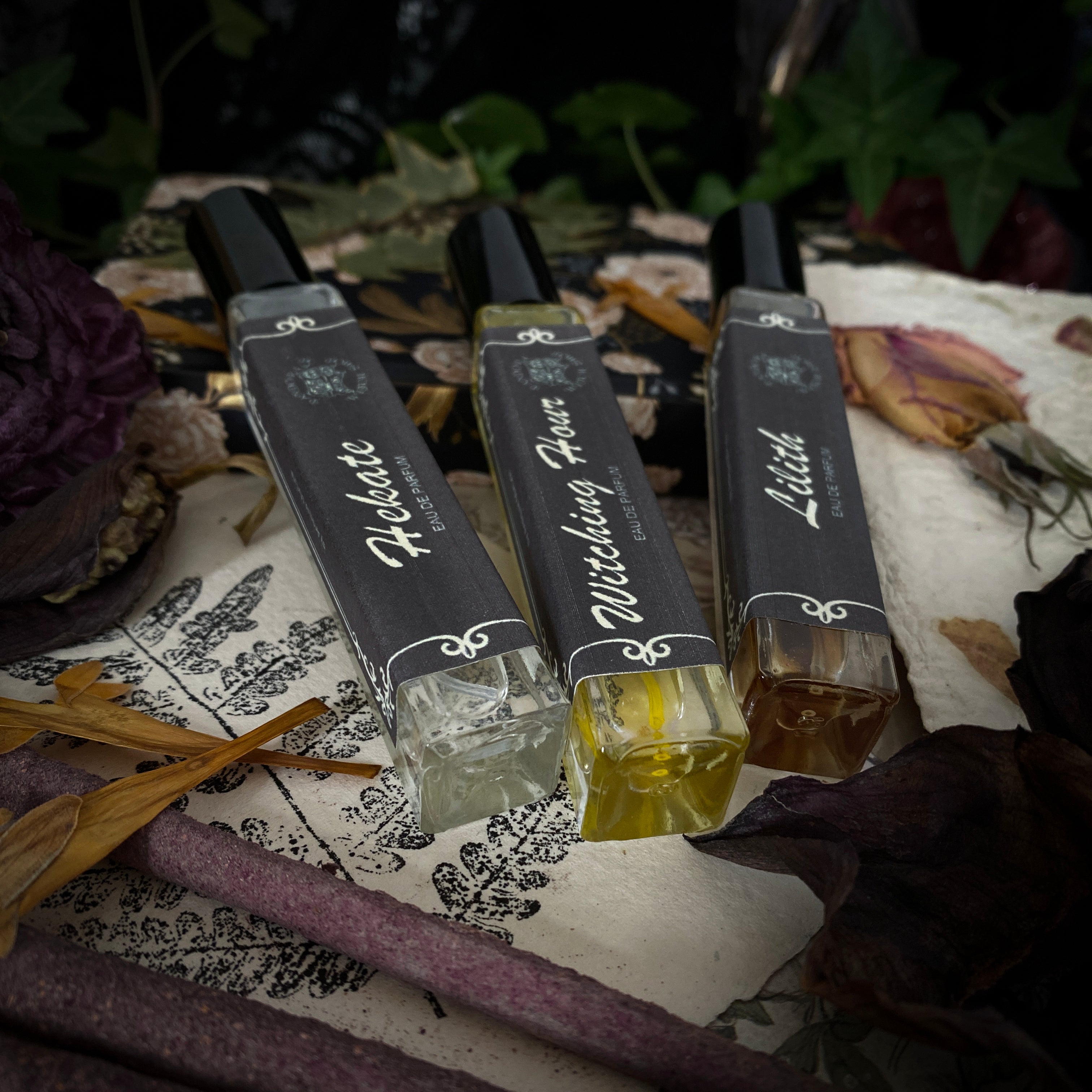 ritual perfumes labeled Lilith, Witching Hour, and Hekate. Styled with dried flowers, candles, and witchcraft accessories on a dark mystical background. Product photo for a witch shop with international shipping
