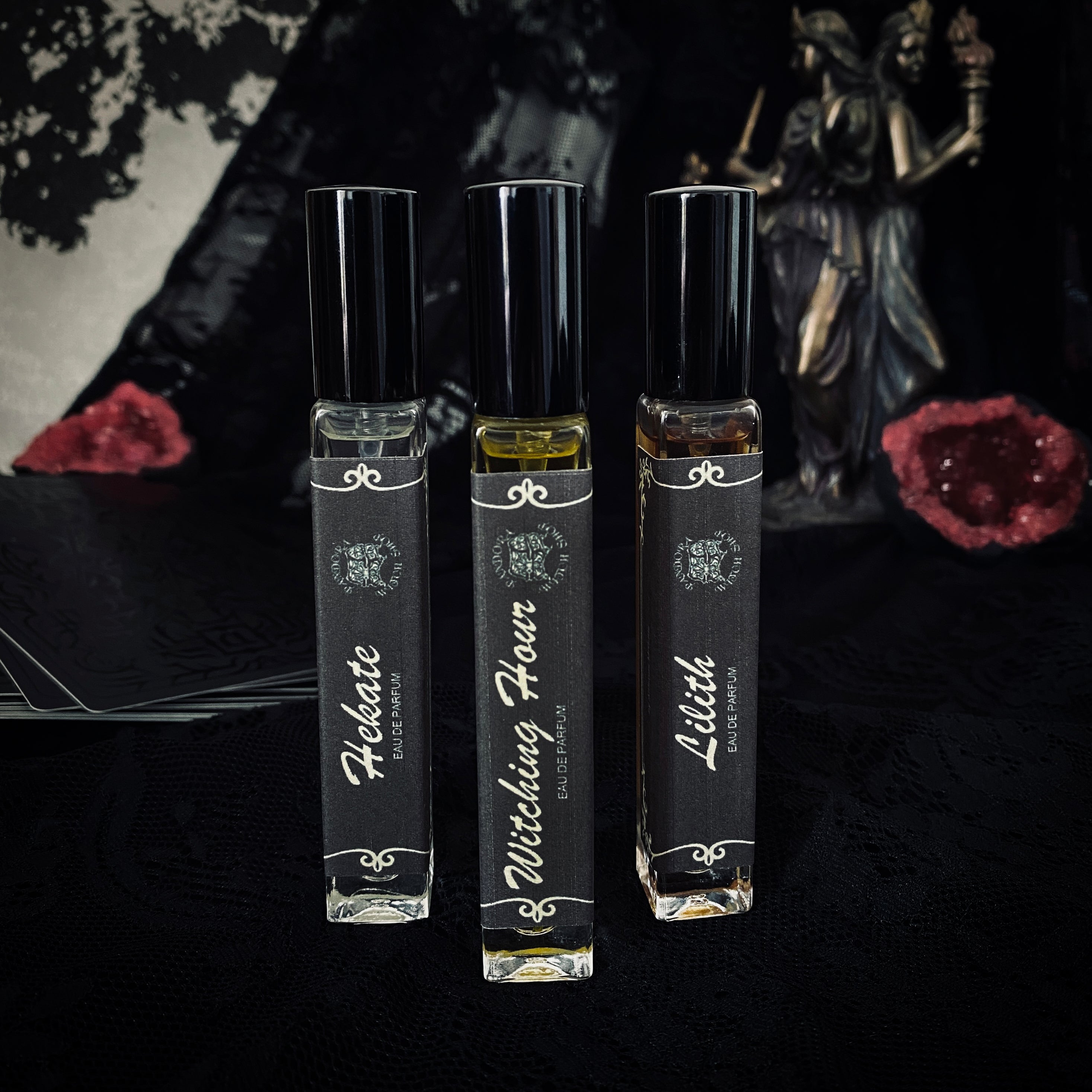 Three black glass bottles of ritual perfumes labeled Lilith, Witching Hour, and Hekate. Styled with dried flowers, candles, and witchcraft accessories on a dark mystical background. Product photo for a witch shop with international shipping