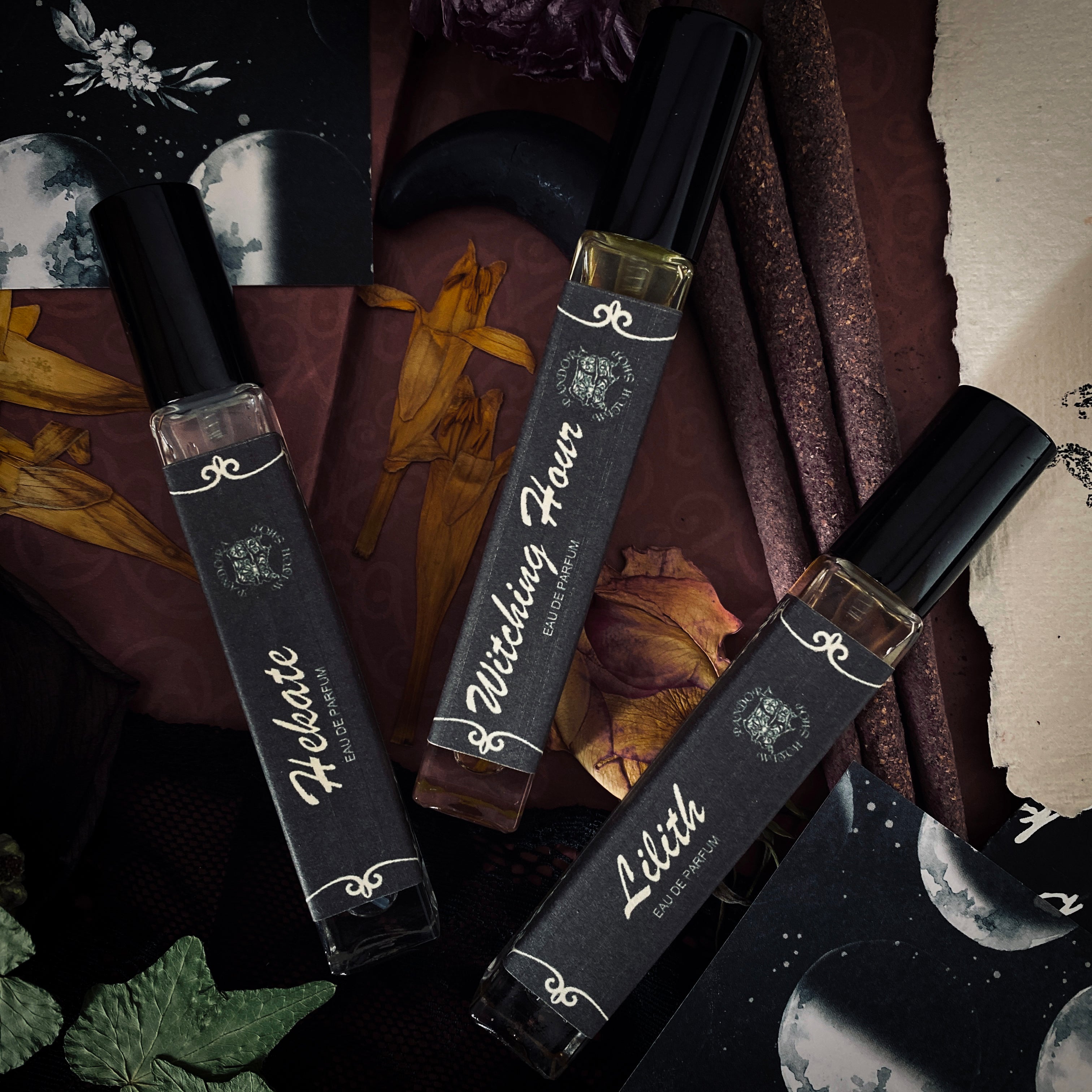 Three black glass bottles of ritual perfumes labeled Lilith, Witching Hour, and Hekate. Styled with dried flowers, candles, and witchcraft accessories on a dark mystical background. Product photo for a witch shop with international shipping