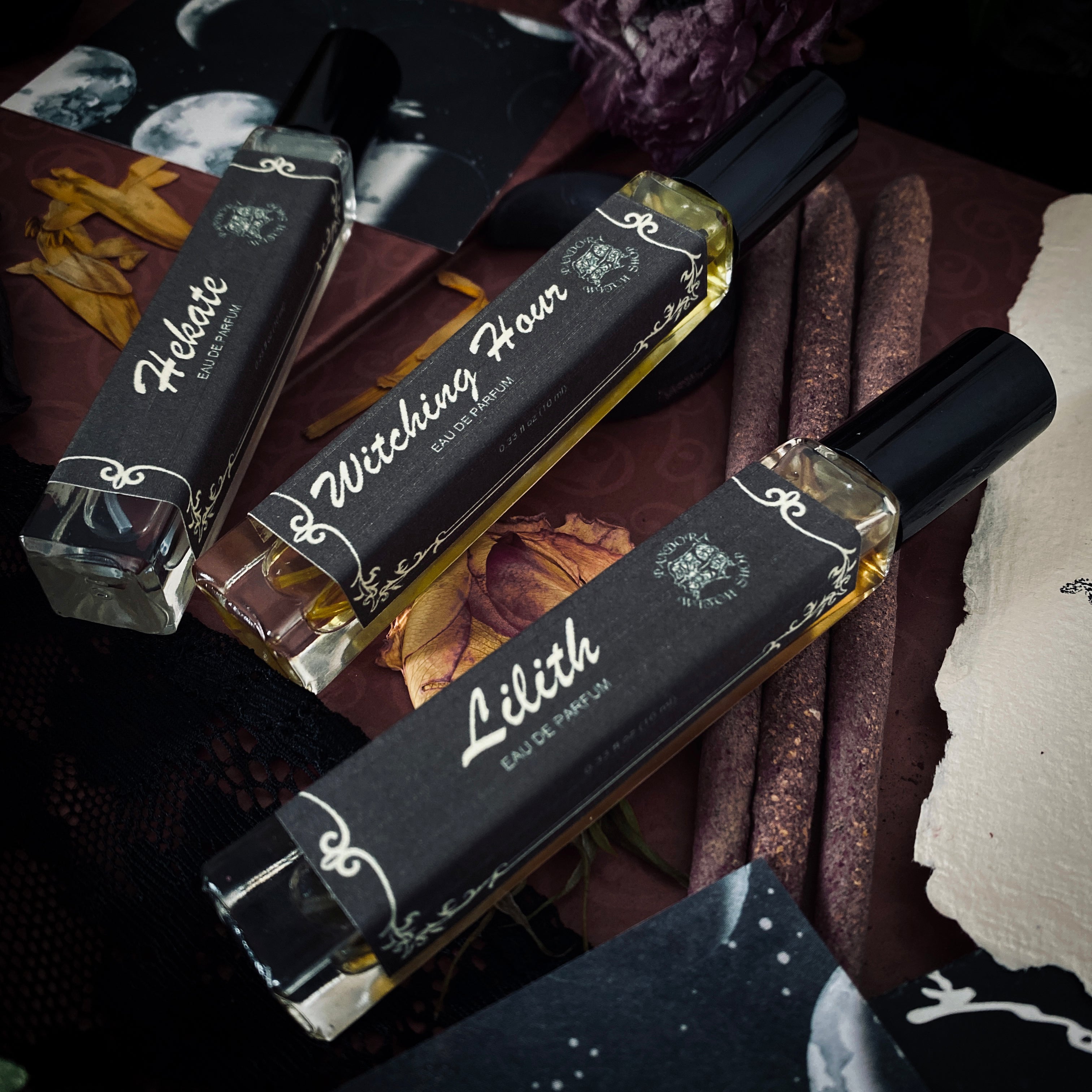 Three black glass bottles of ritual perfumes labeled Lilith, Witching Hour, and Hekate. Styled with dried flowers, candles, and witchcraft accessories on a dark mystical background. Product photo for a witch shop with international shipping